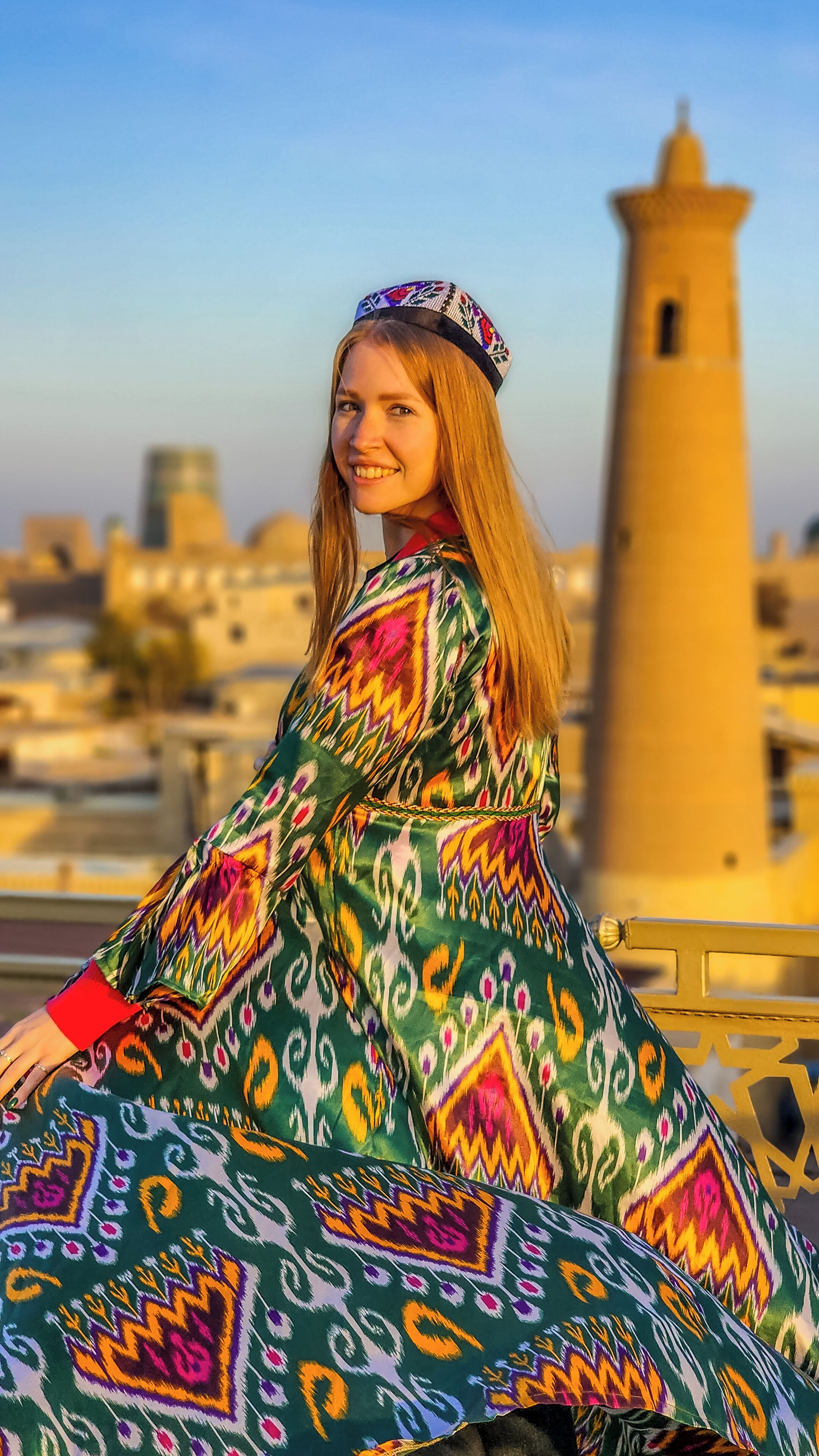 A person in a colorful traditional dress sitting on a bench with a cityscape and a tower in the background.
