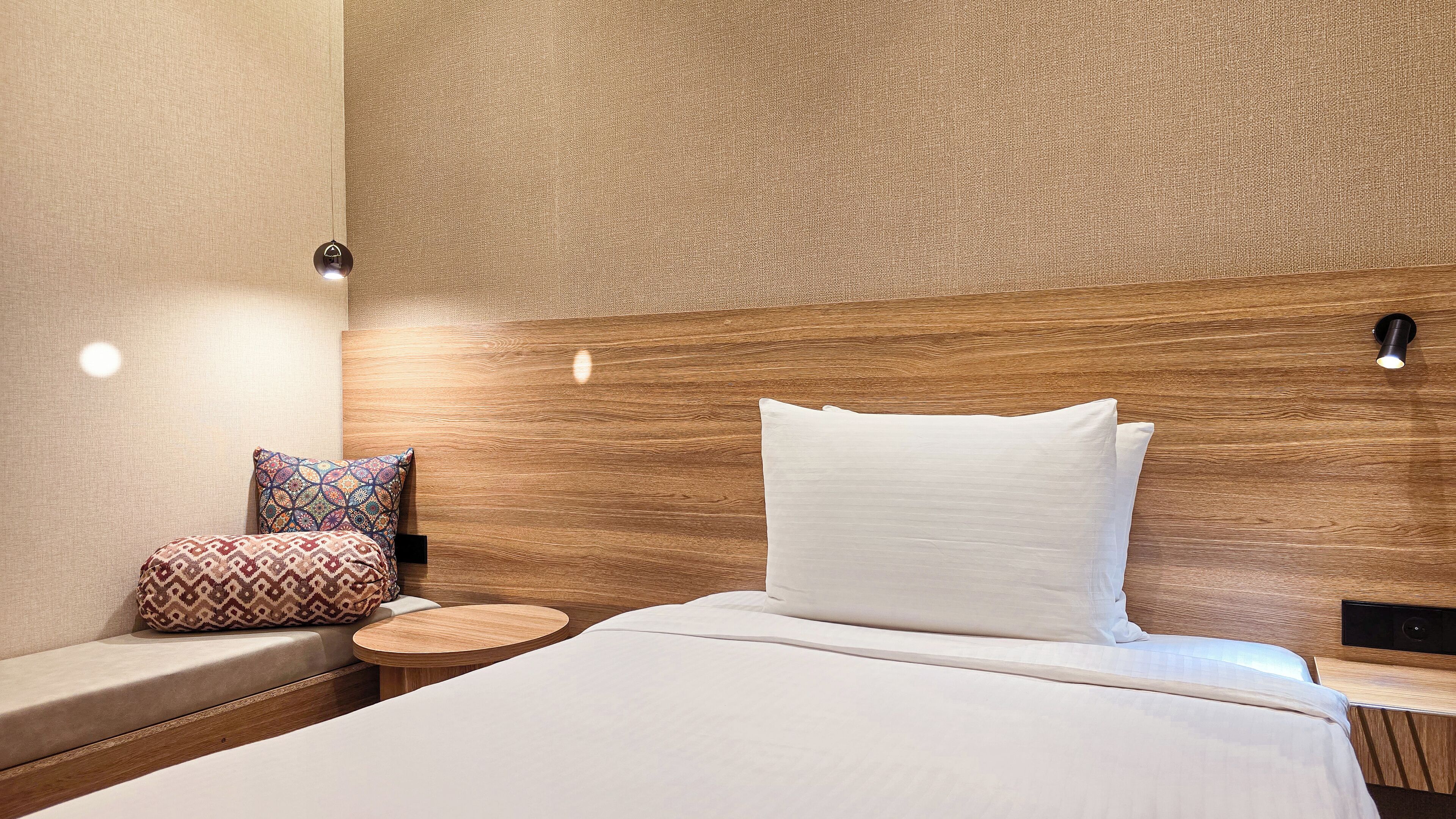 Habitación de hotel con cabecera de madera, cama con ropa blanca, mesita de madera redonda y almohada decorativa estampada.