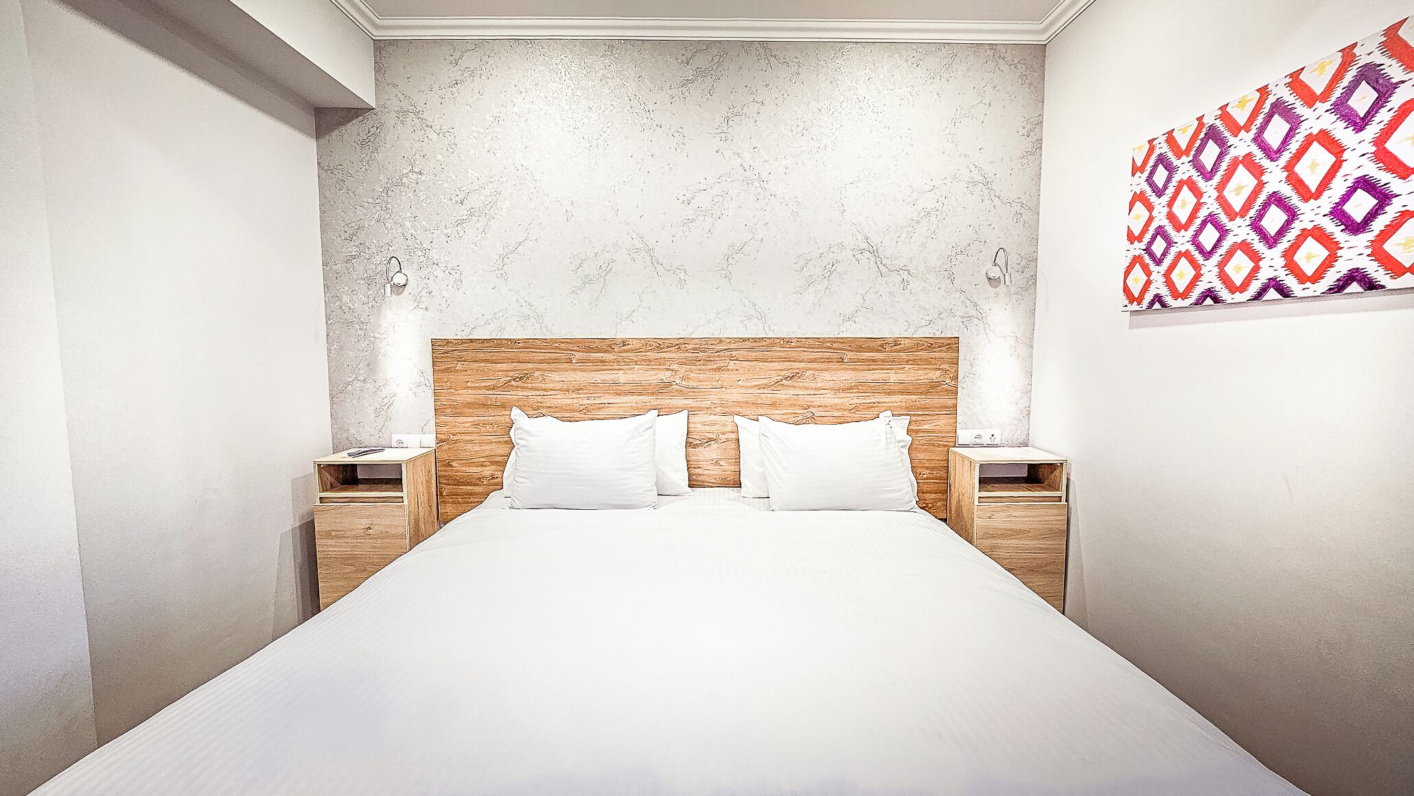 Habitació d'hotel moderna amb un llit gran, capçalera de fusta, roba de llit blanca i dues taules de nit amb làmpades.