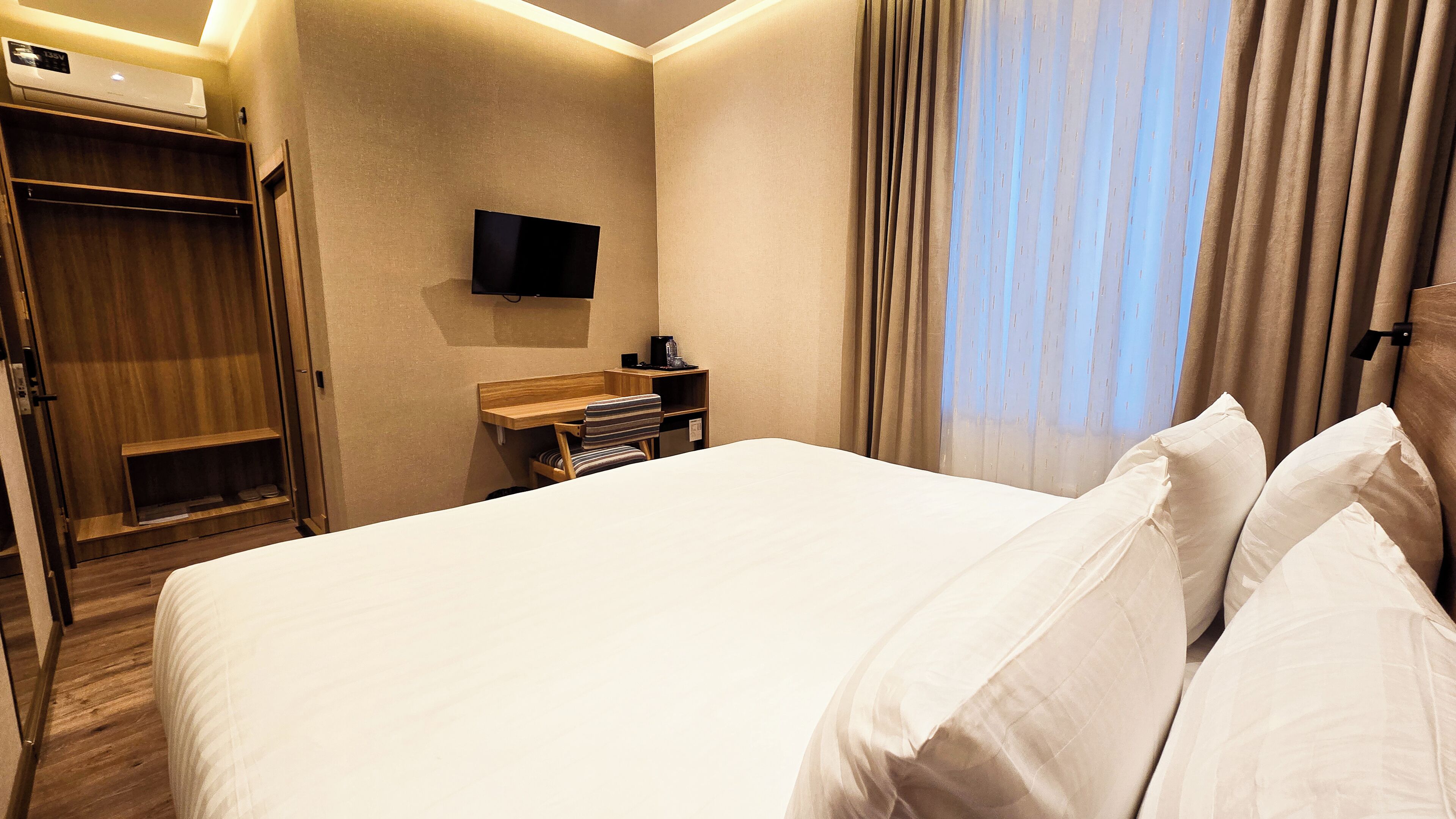 A hotel room with a large bed, a wooden desk, a television mounted on the wall, and a wardrobe.