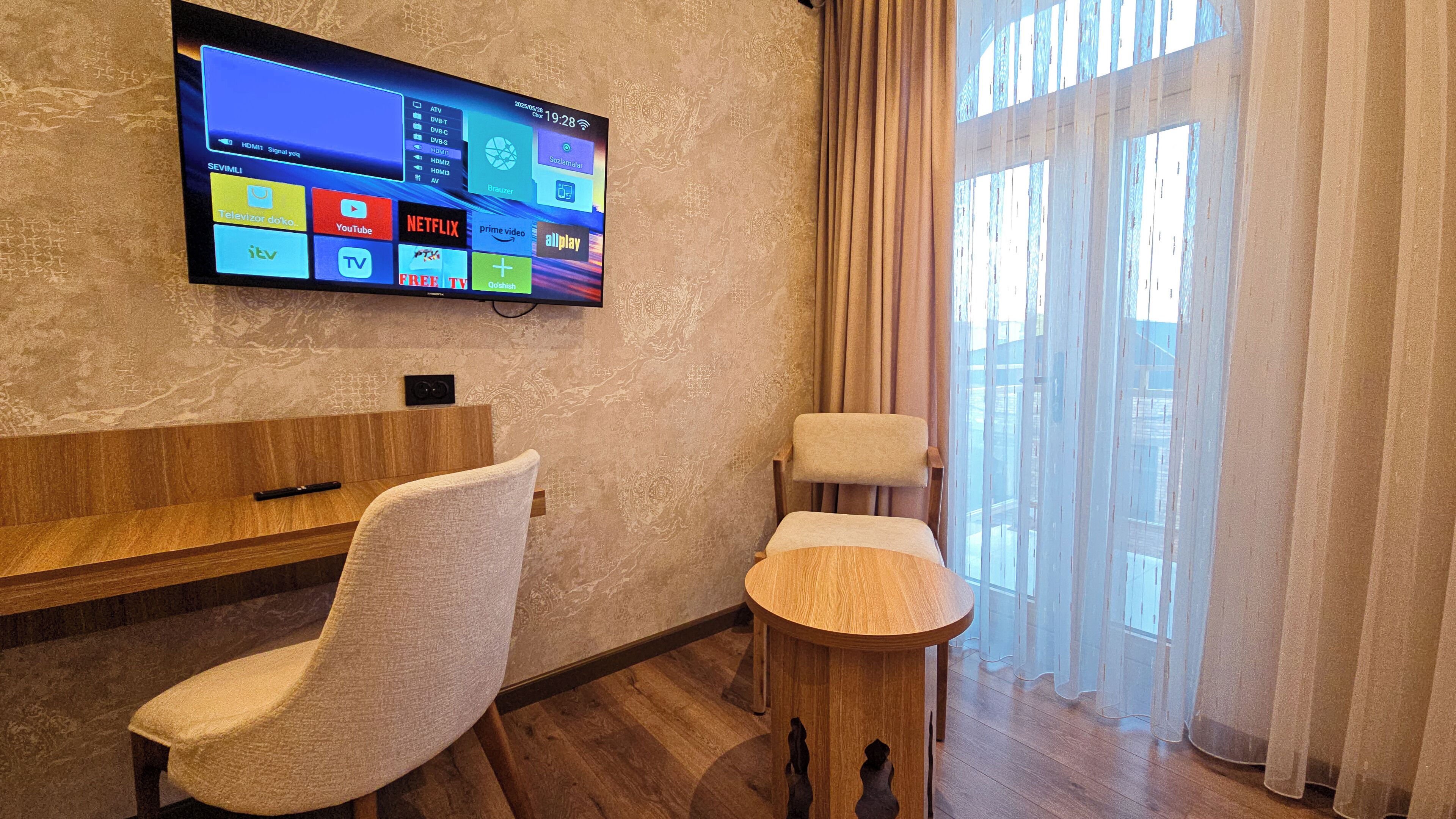 A hotel room with a large flat-screen TV displaying various streaming services, a wooden bench, a cushioned chair, a small wooden table, and a large window with sheer curtains.