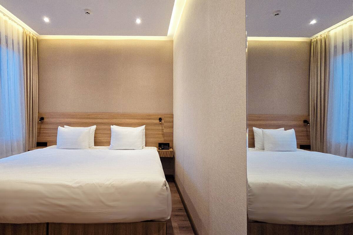 Ein Hotelzimmer mit zwei Betten, beigen Wänden, Holz-Kopfteilen und einem großen Fenster mit Vorhängen.