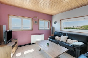 A living room with a television on a wooden stand, a black leather sofa, a wooden coffee table, and a view of the sea through large windows. - Holiday Home 'Villa Timao Plage' with Sea View, Private Terrace and Wi-Fi (Batz-sur-Mer)