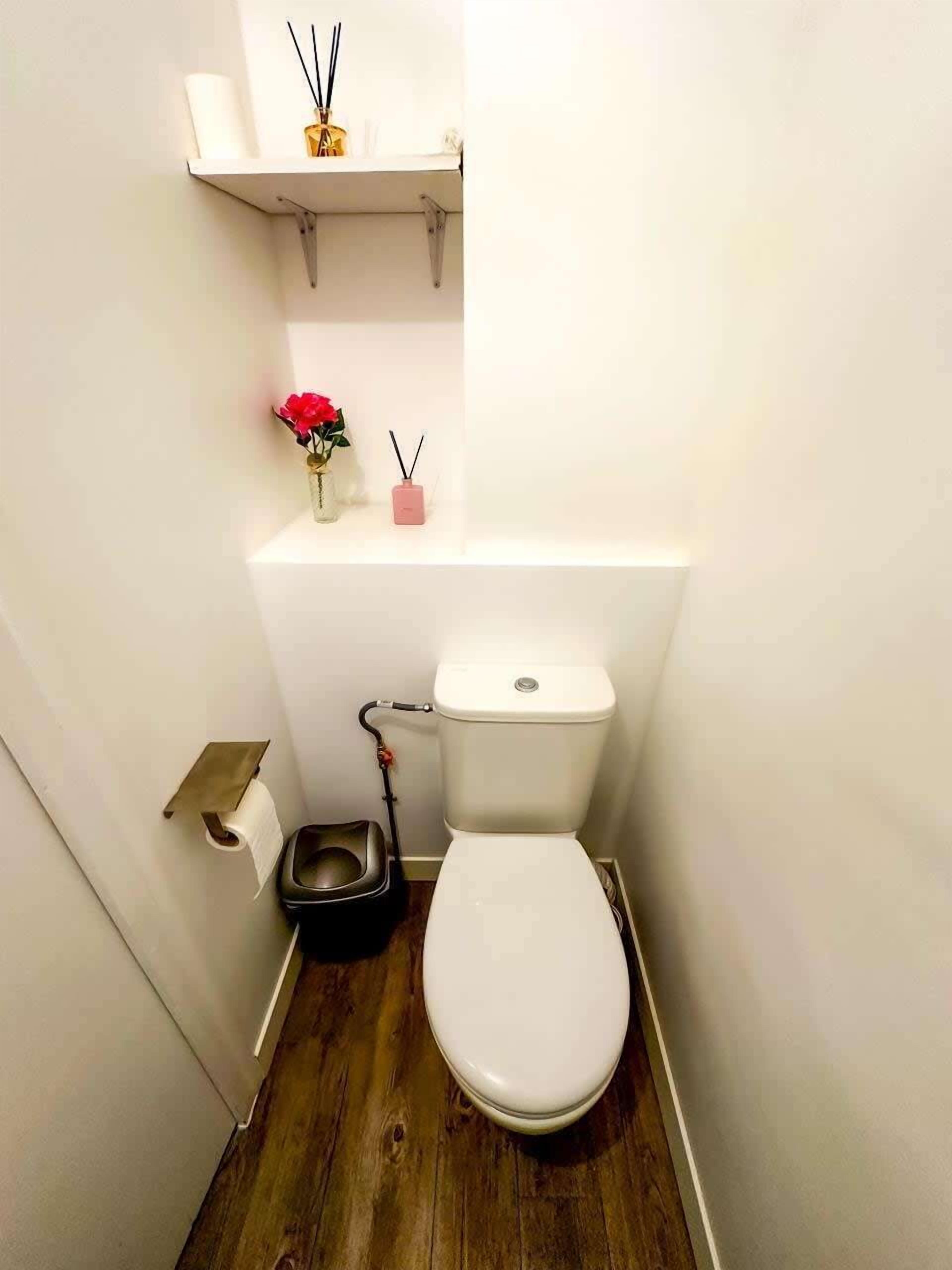 A small bathroom with a toilet, a bidet, a toilet paper holder, and a shelf with a vase and a small pink container.