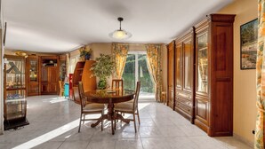 Un coin repas avec une table ronde, des chaises et un luminaire au plafond. Il y a une fenêtre avec des rideaux et une vue sur la verdure à l’extérieur.