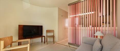 A modern living room with a flat-screen TV, wooden furniture, a grey sofa, and a pink and white striped partition.