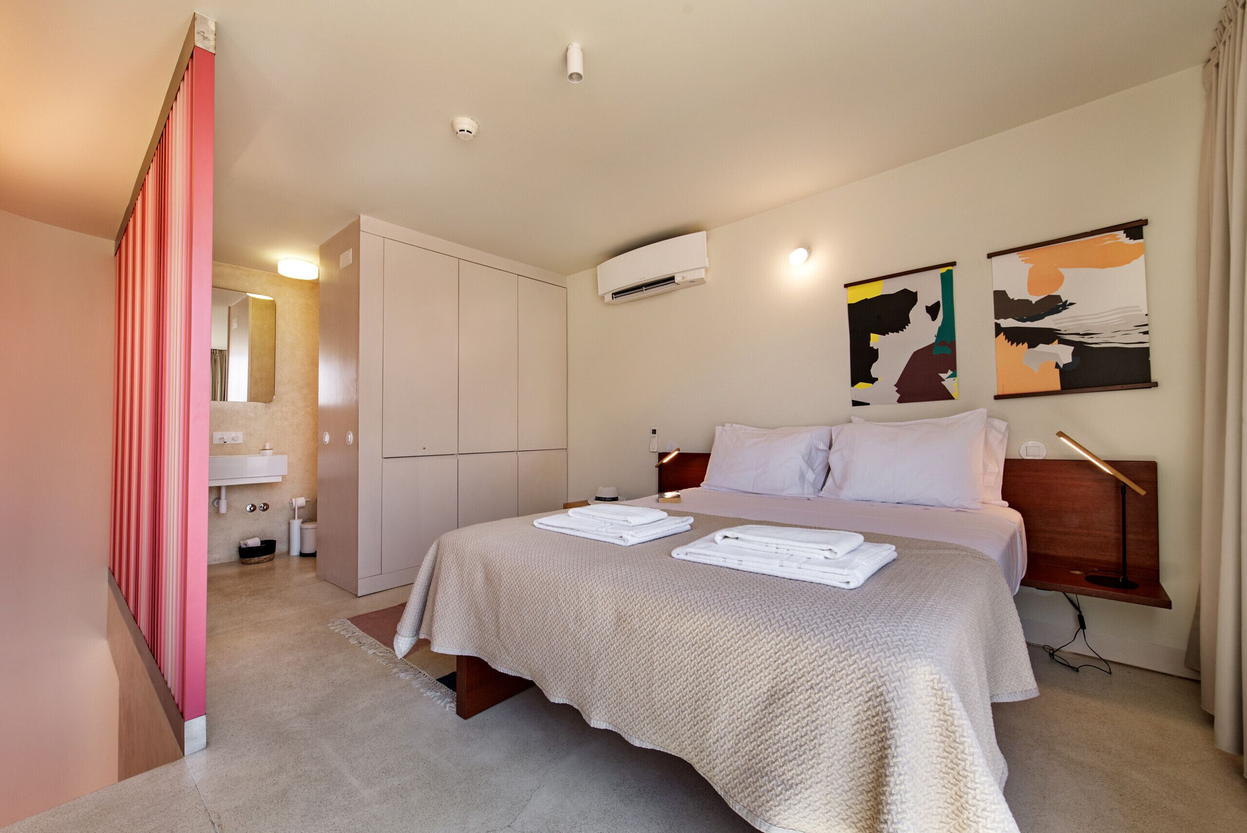 Una habitación de hotel con cama, mesita de noche, armario, espejo, unidad de aire acondicionado y cuadros en la pared.