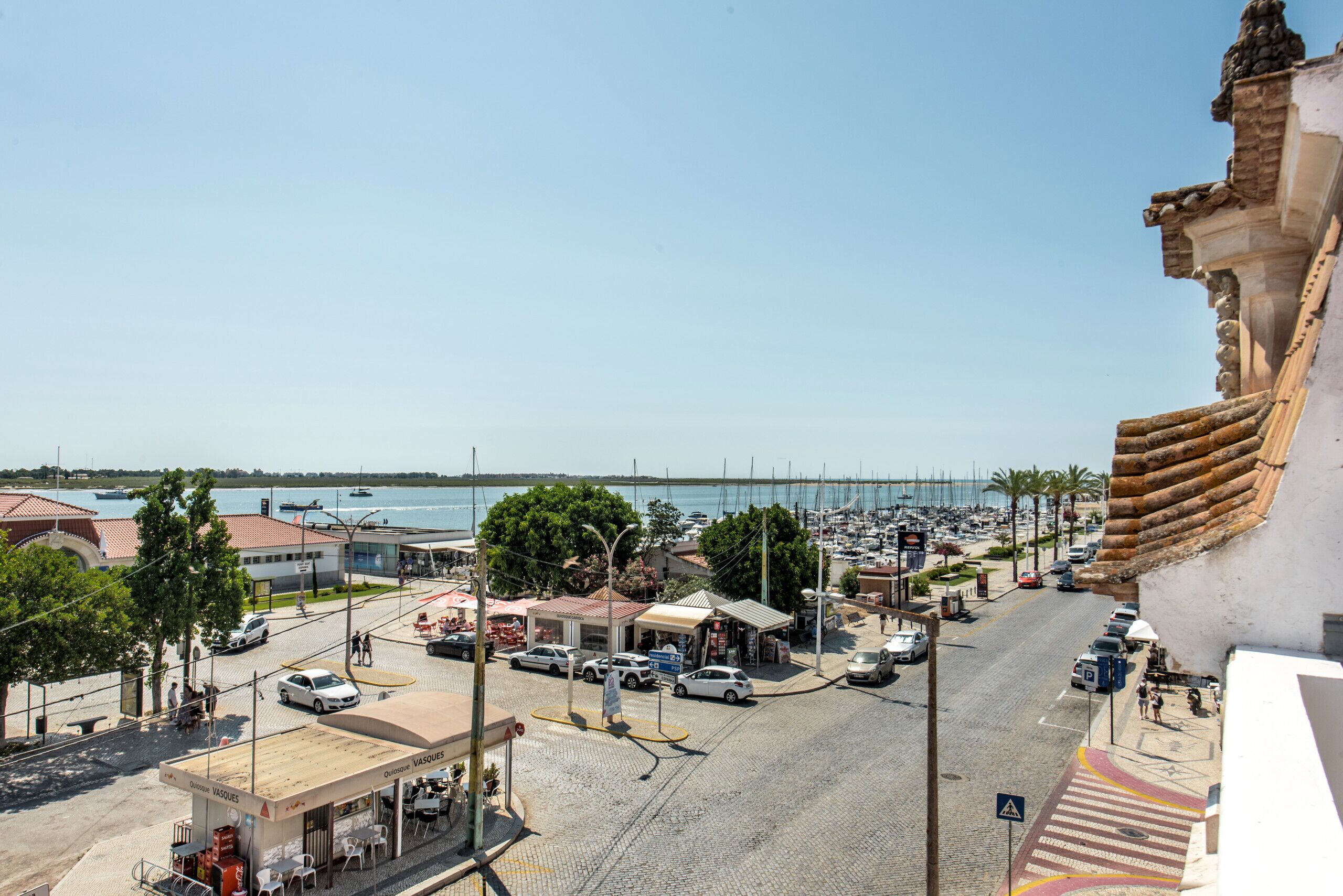Une rue côtière bordée d’immeubles, avec un port de plaisance et un pont en arrière-plan.