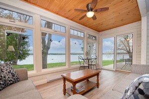 A spacious room with large windows overlooking a lake, a ceiling fan, and a wooden table. - Waterfront Cottage - Lake and Loon- Family Friendly Accessible (Buckhorn)