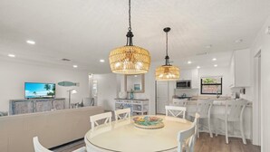 Eine moderne Küche mit weißen Schränken, ein Essbereich mit rundem Tisch und weißen Stühlen sowie ein Wohnbereich mit Sofa und Fernseher.