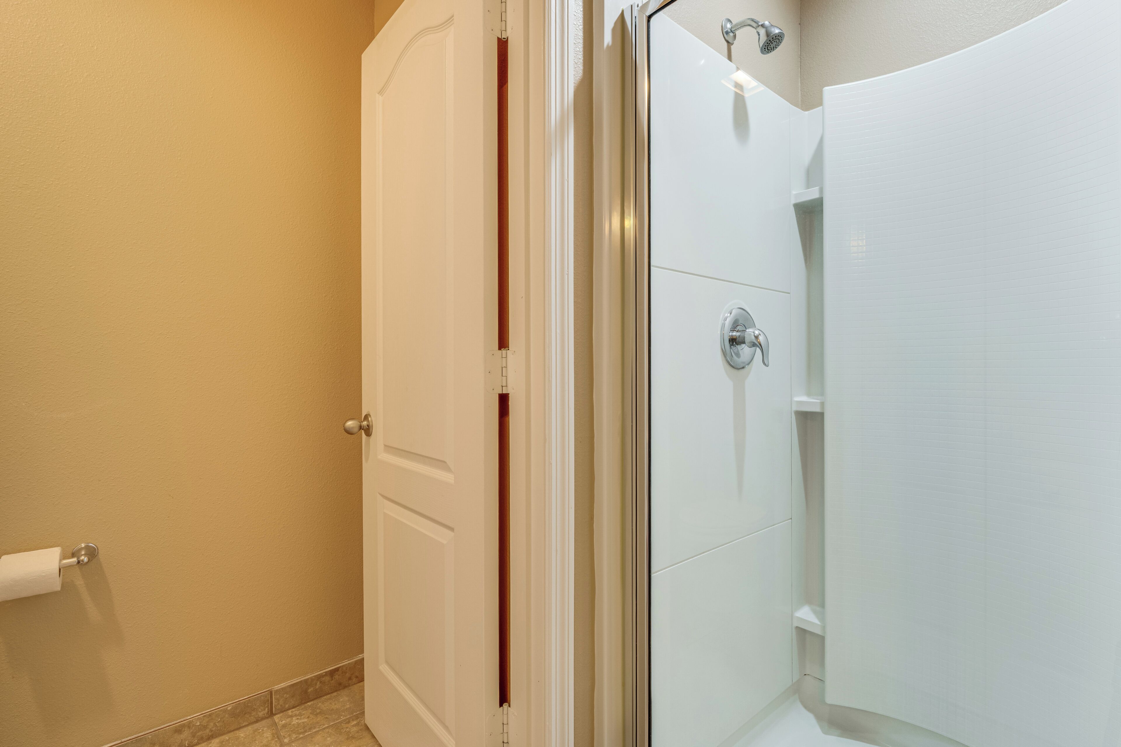 A bathroom with a glass shower enclosure, a toilet with a paper towel holder, and a door with a vertical handle.
