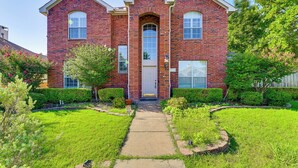A two-story brick house with a central entrance, a front yard with a pathway, and a small garden area. - DFW Sports Hub: Home w/ Pool & Hot Tub in Frisco! (Frisco)