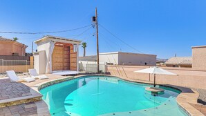 Una piscina con borde curvo, rodeada de un patio de ladrillo y un área de descanso cubierta con sillones blancos y una sombrilla.