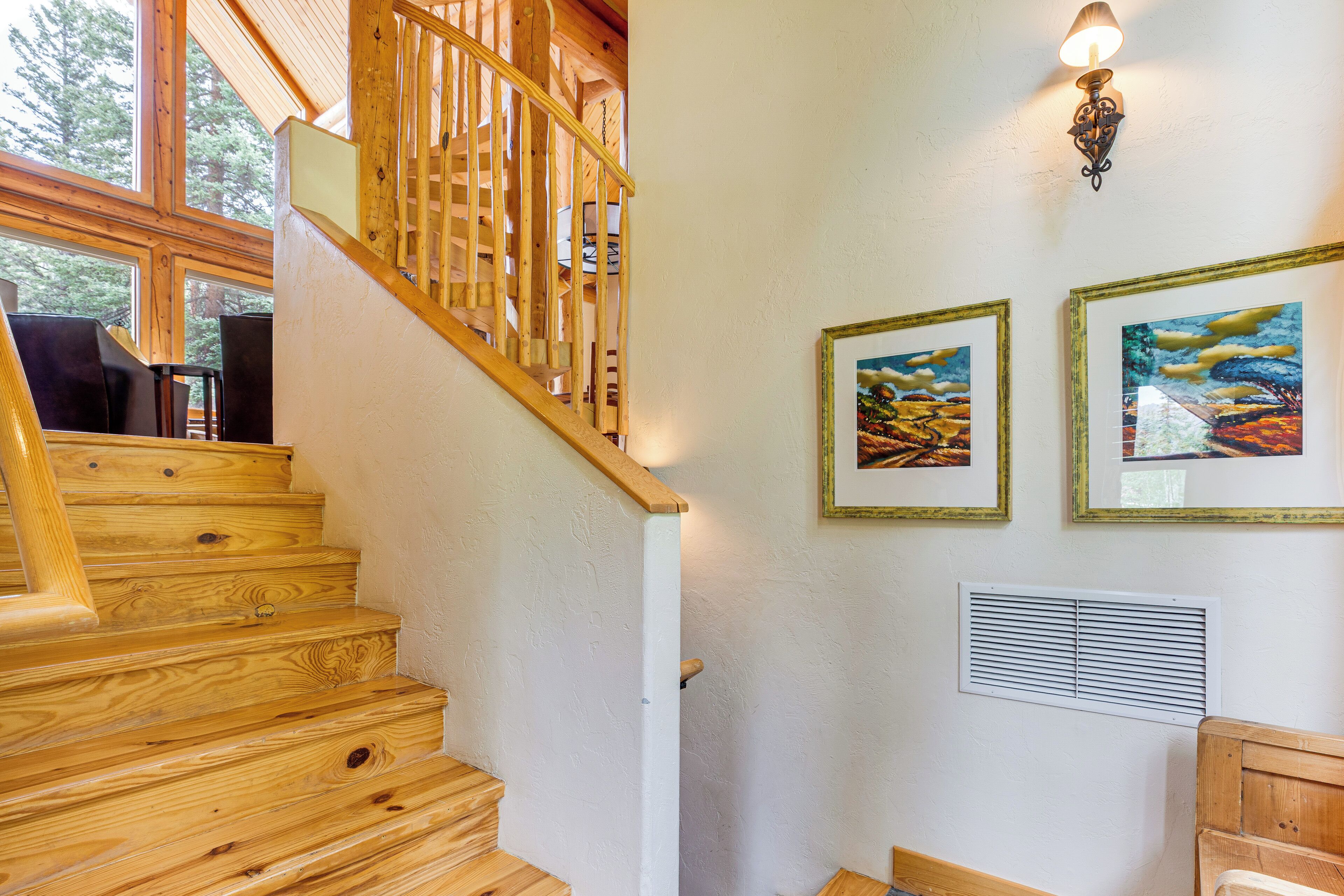 A wooden staircase with a railing, a wall-mounted air vent, and two framed landscape paintings.