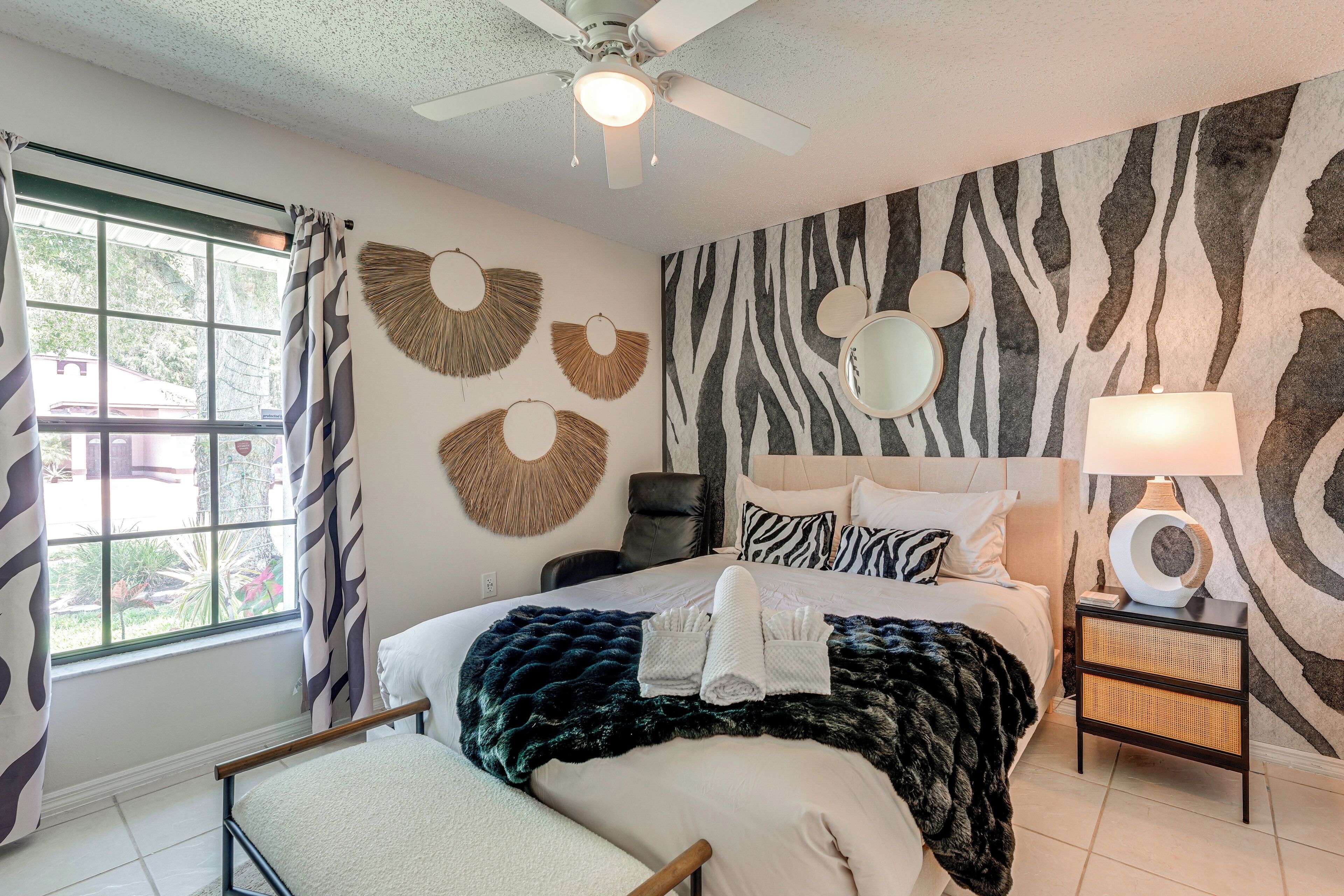 A bedroom with a zebra-patterned wall, a ceiling fan, a bed with a dark blanket, a bedside table with a lamp, and a window with curtains.