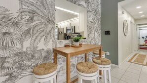 A dining area with a wooden table and white stools, a kitchen in the background, and a tropical wallpaper design. - Disney Getaway w/ Private Hot Tub 14 Mi to Parks (Kissimmee)