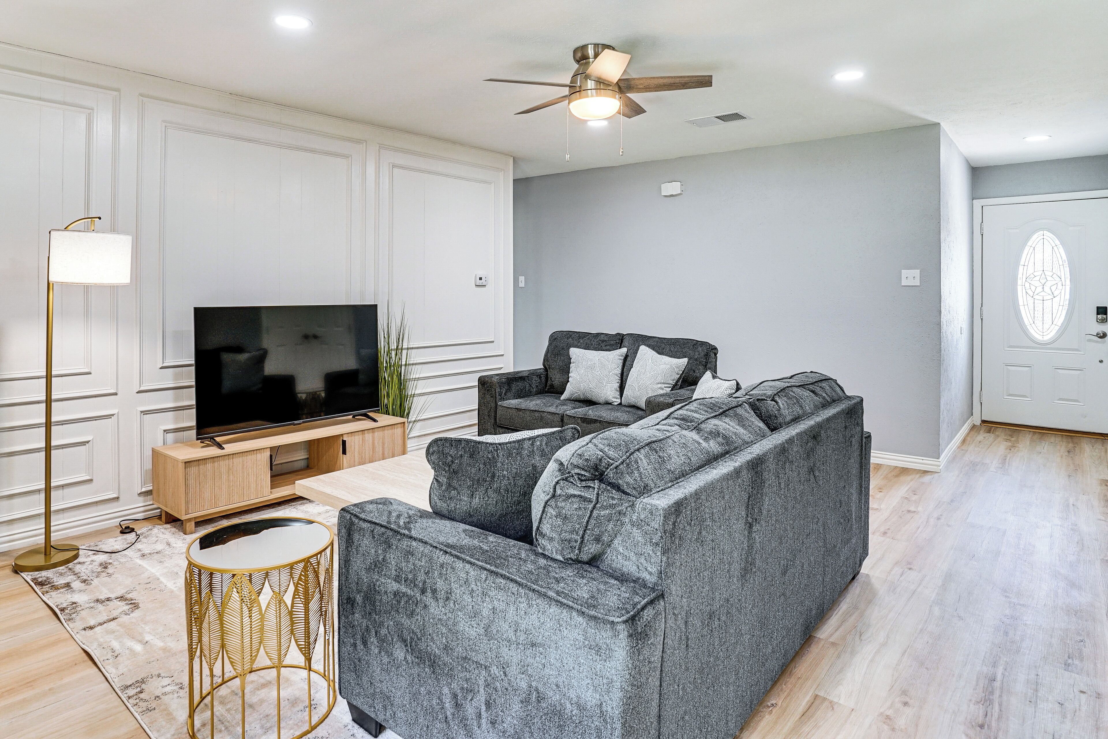 Ein modernes Wohnzimmer mit einem grauen Sofa, einem Fernseher auf einem Holzboden, einer Stehlampe und einem Deckenventilator.