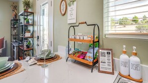 A kitchen with a dining area, a window with blinds, a shelf with plants, a sign with a message, and a rack with food items.