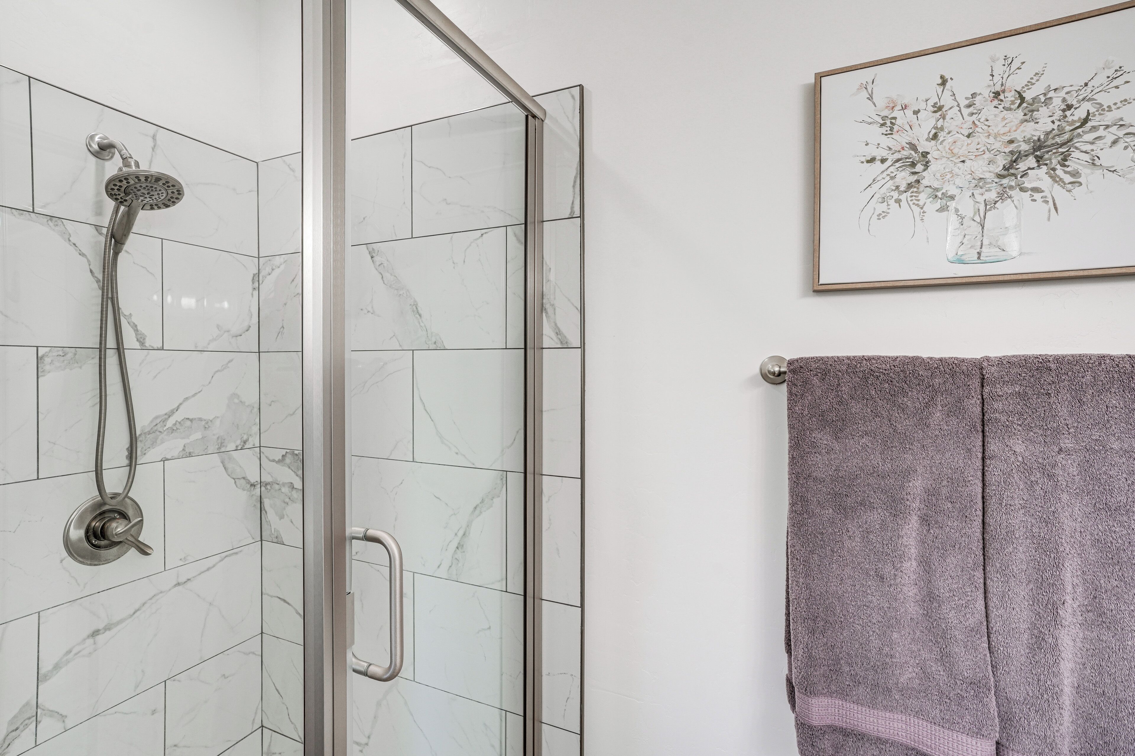 A shower with a handheld showerhead, a glass door, and a marble-tiled wall.