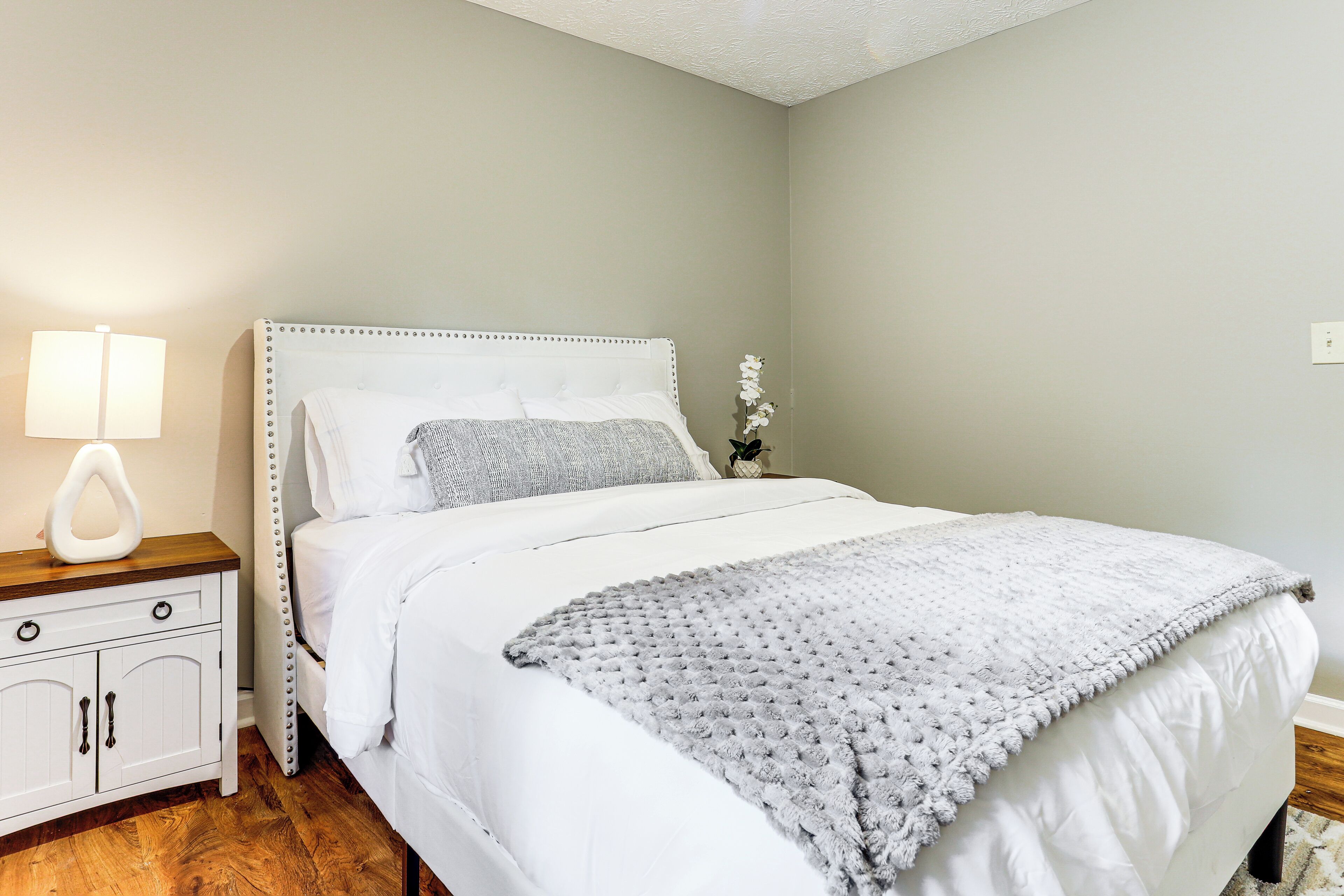 Un letto rifatto con un poggiatesta bianco, una coperta grigia e un comodino con una lampada.