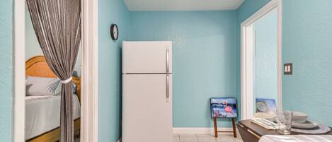 A compact room with a bed, a white refrigerator, a small table with a painting, and a mirror on the wall.