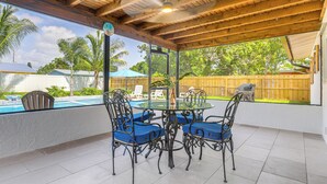 Eine überdachte Terrasse mit Glastisch und -stühlen, einem Deckenventilator und Blick auf einen Pool und Garten.
