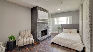 Une chambre avec une cheminée en briques, un fauteuil à motifs, un grand lit et une vue sur l’extérieur.