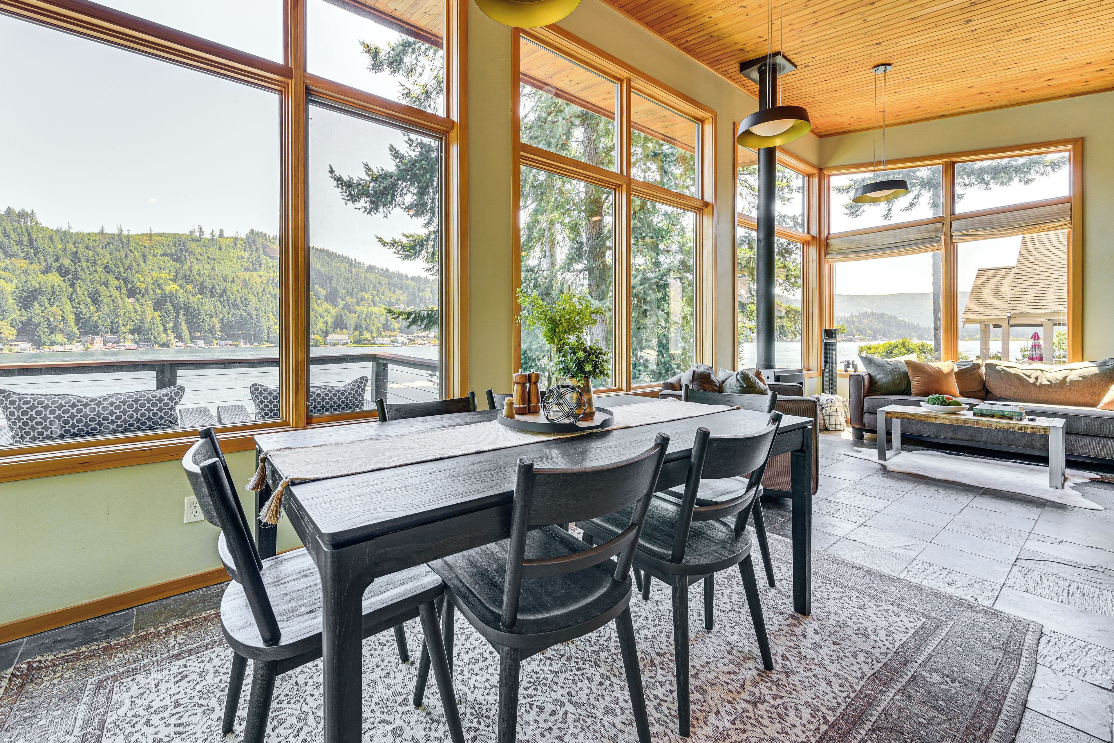 Une pièce spacieuse dotée de grandes fenêtres offrant une vue sur une colline boisée. On peut apercevoir un coin repas avec une table et des chaises en bois, ainsi qu’un coin salon avec un canapé et une table basse.