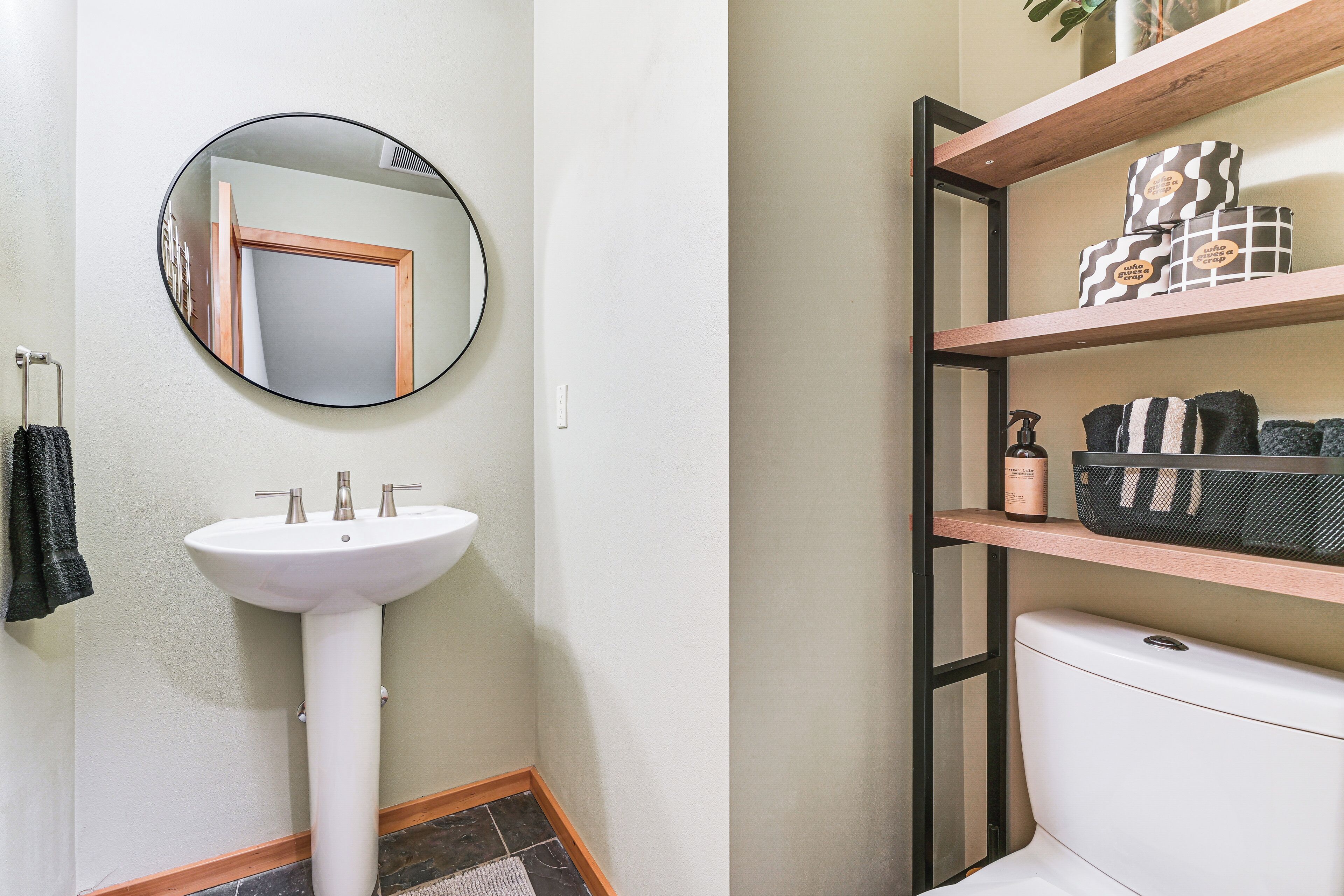 A bathroom with a white pedestal sink, a round mirror, a toilet, and a shelf with toiletries.