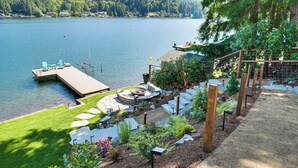A lakeside deck with a hot tub, seating area, and a dock extending into the water. - Modern Summit Lake Escape - Spacious Deck w/ View! (Olympia)