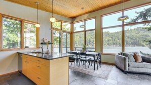 A modern kitchen with a large island, wooden ceiling, and large windows offering a view of a forested area. - Modern Summit Lake Escape - Spacious Deck w/ View! (Olympia)