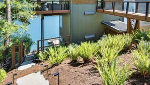 A multi-level wooden structure with a balcony overlooking a body of water, surrounded by ferns and a stone pathway. - Modern Summit Lake Escape - Spacious Deck w/ View! (Olympia)