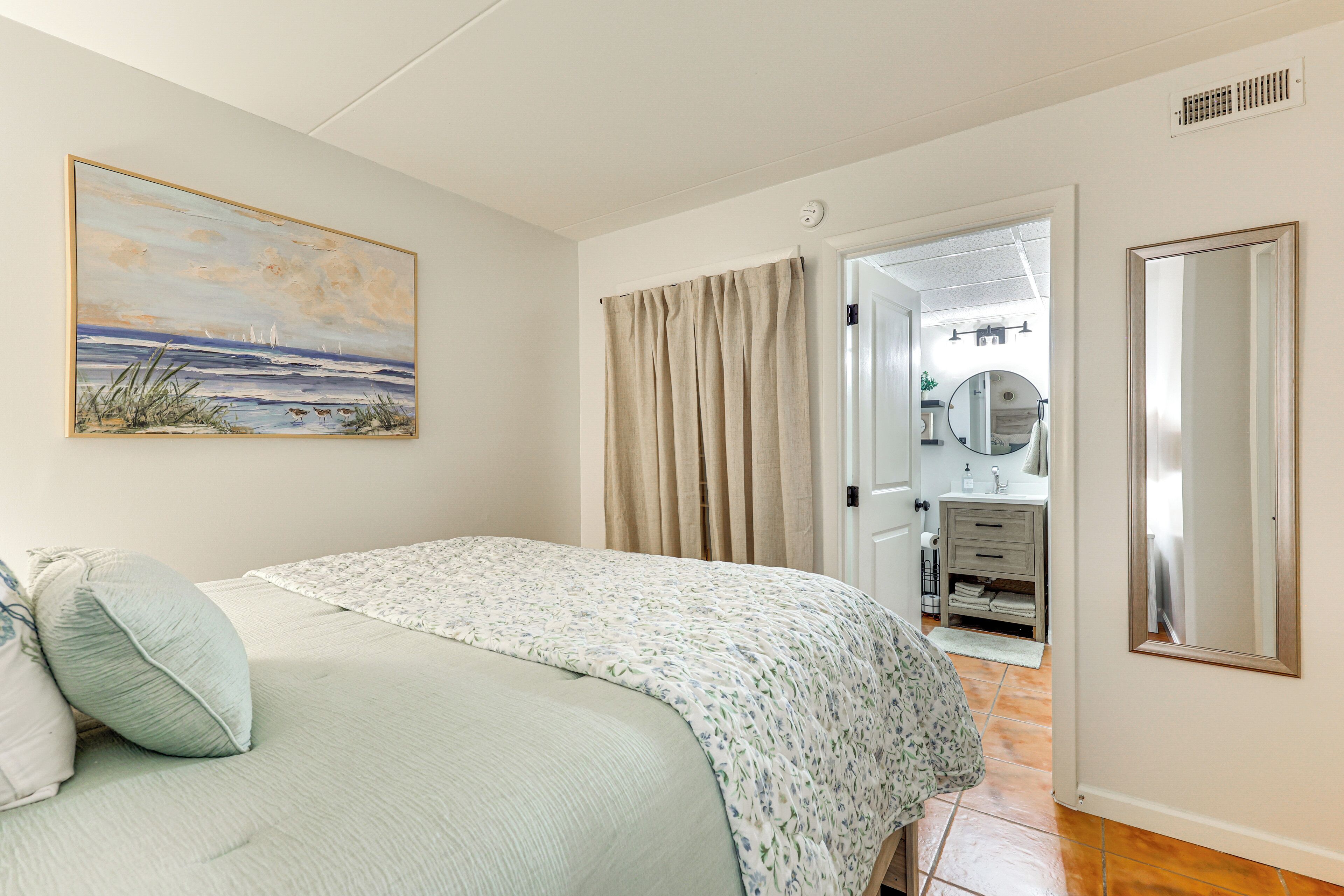 A bedroom with a bed, a painting, a bathroom with a mirror, and a door leading to another room.