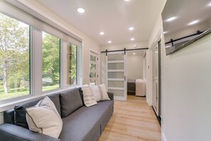 A modern living room with a grey sofa, large windows, a wooden floor, and a sliding glass door leading to a closet. - Container Cottage Between Knoxville & Sevierville! (Knoxville)