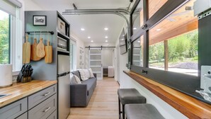 A compact kitchen with a refrigerator, wooden countertop, and hanging utensils. A gray sofa and bench are in the dining area, with a window offering a view of greenery.