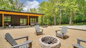 A modern outdoor seating area with Adirondack chairs and a stone fire pit, surrounded by a gravel patio and a wooden deck with glass doors.