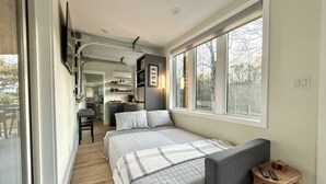 A modern bedroom with a large window, a bed with a grey headboard, and a small wooden stool with remote controls.