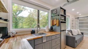 A modern kitchen with a large window, wooden countertop, grey cabinets, and a grey sofa in the adjacent living area.