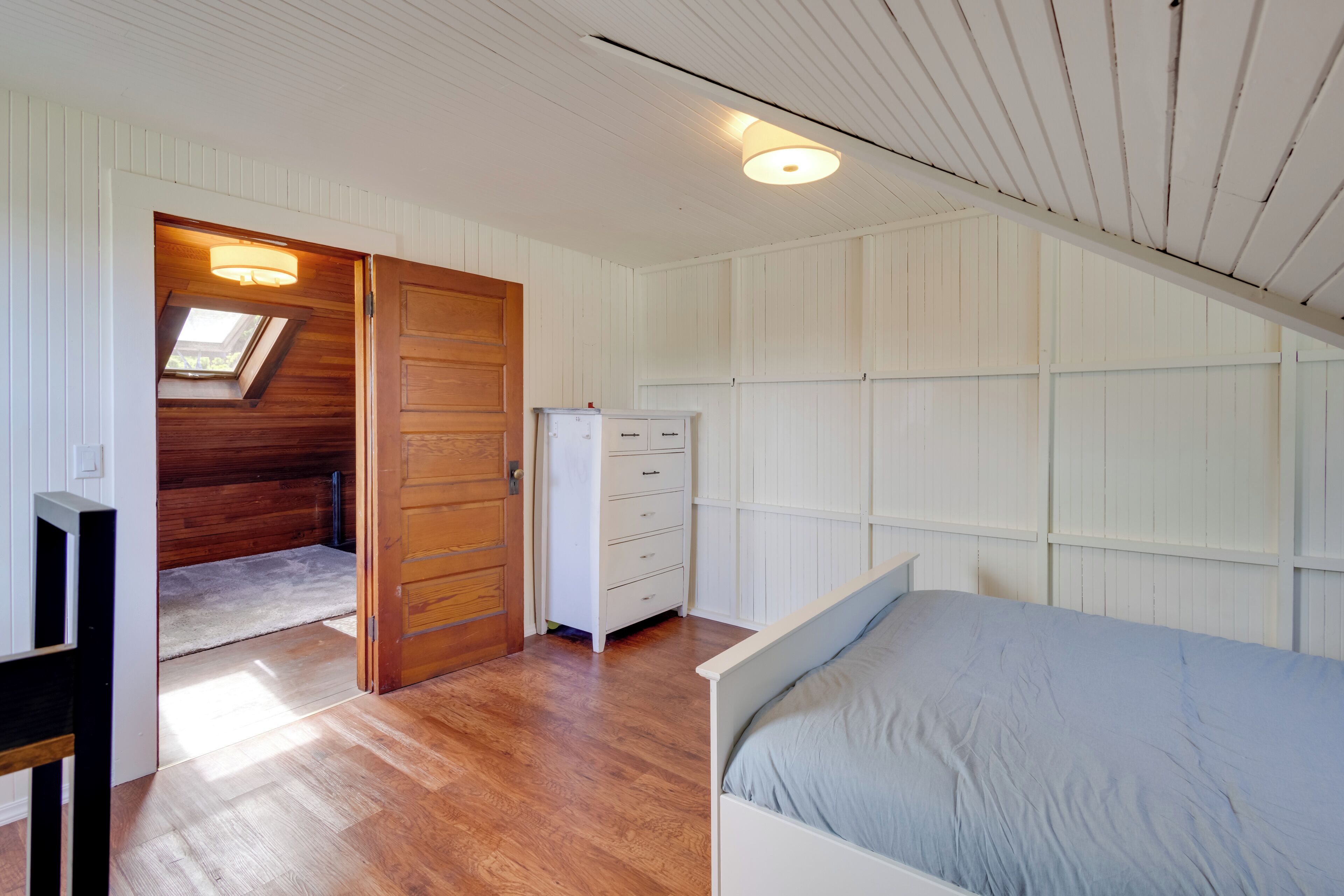 Une chambre avec une porte en bois, une commode blanche, un lit avec un matelas bleu et une lucarne.