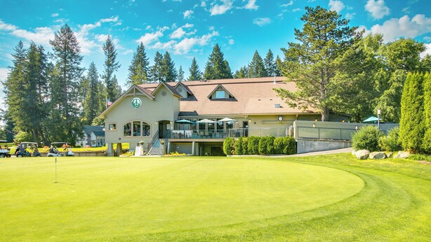 Un campo de golf con un club de campo que cuenta con una amplia entrada con ventanales, asientos al aire libre y un césped bien cuidado.