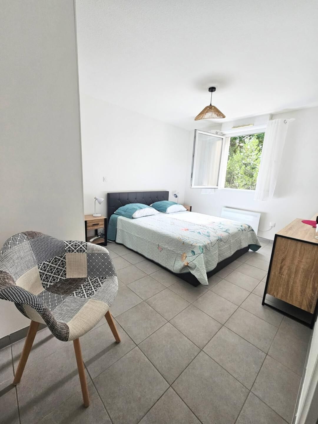 A bedroom with a bed, a chair, a desk, a window, and a hanging light fixture.