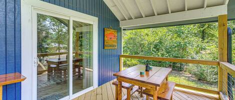 A wooden deck with a table and benches, a glass door leading to an outdoor area, and a view of greenery through the windows.