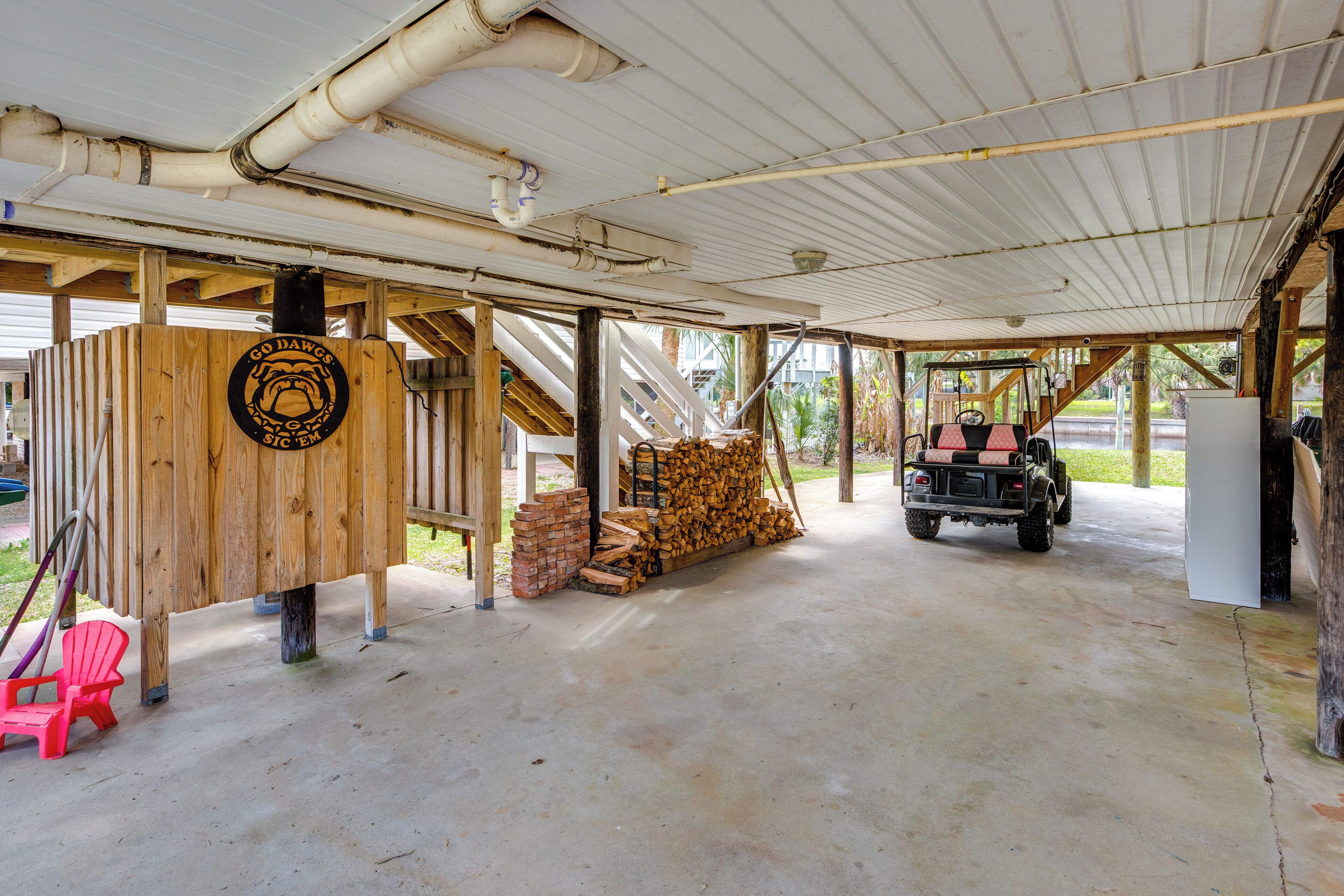 A covered outdoor area with a wooden structure, a stack of firewood, a vehicle, and a red chair.
