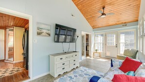 A spacious living room with a wooden ceiling, a large flat-screen TV mounted on the wall, a white dresser, a patterned rug, and a bed with colorful pillows. - Fish, Swim & Boat: Colonial Beach Bungalow (Colonial Beach)