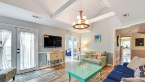 A spacious living room with a blue sofa, a green armchair, and a turquoise coffee table. There is a television mounted on the wall and a chandelier hanging from the ceiling. - Fish, Swim & Boat: Colonial Beach Bungalow (Colonial Beach)