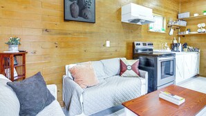 A cozy living room with a wooden floor, a sofa with cushions, a coffee table, a kitchen area with a stove and shelves, and a wall-mounted air conditioner.