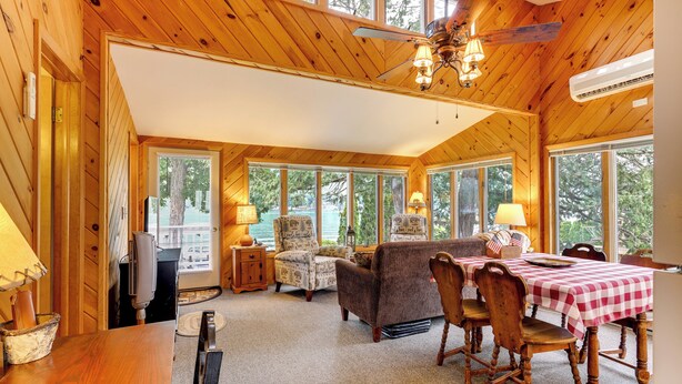 A wooden cabin interior with a dining area, living room, and large windows. - Waterfront Cottage on Lake George w/ 2 Docks! (Queensbury)