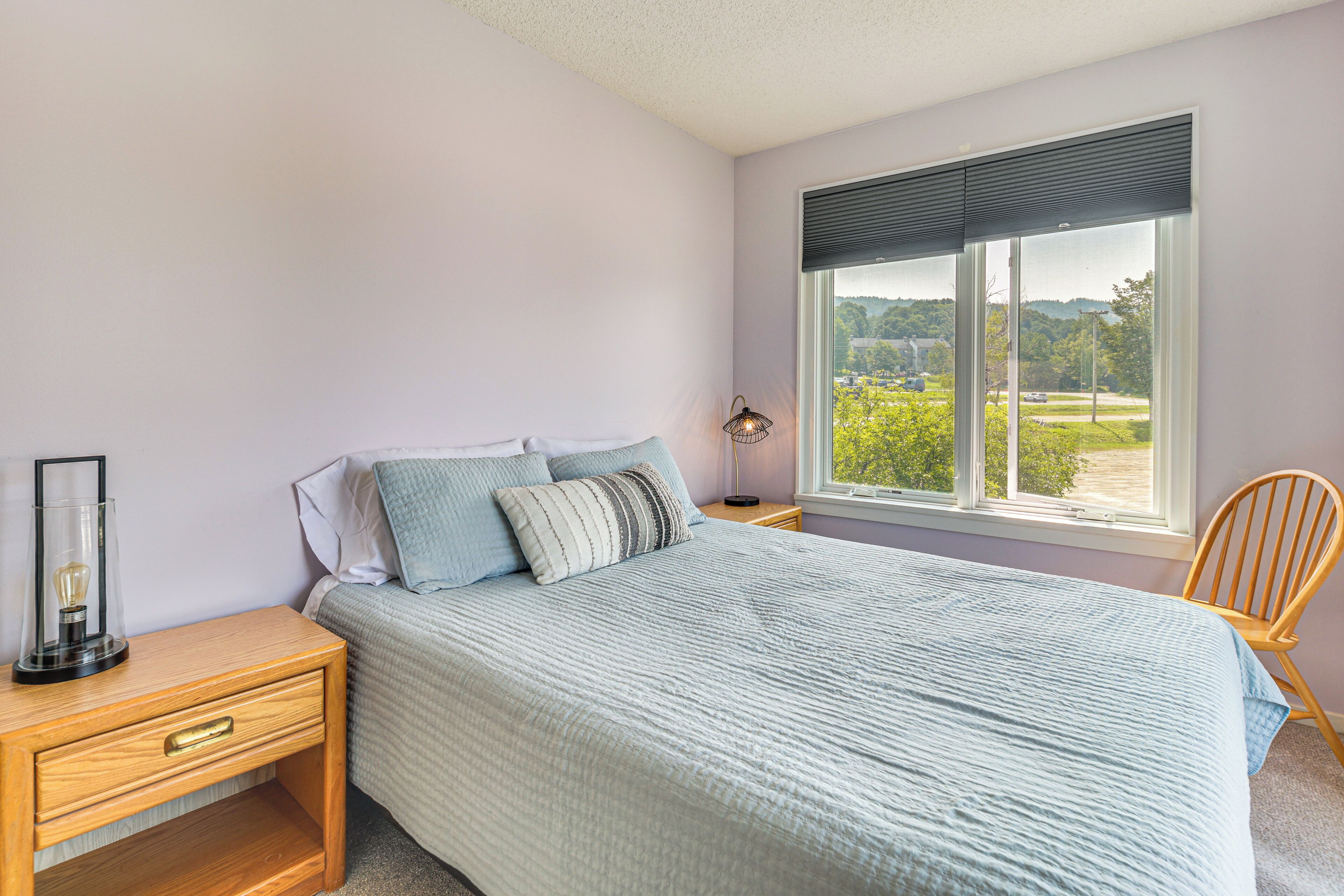 A bedroom with a bed, a nightstand, a chair, and a window with a view of greenery.