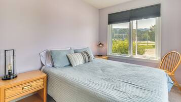 A bedroom with a bed, a nightstand, a chair, and a window with a view of greenery.