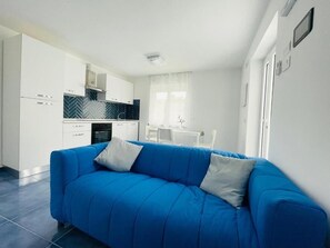 Una cuina moderna amb armaris blancs, un sofà blau i una zona de menjador amb taula i cadires blanques.